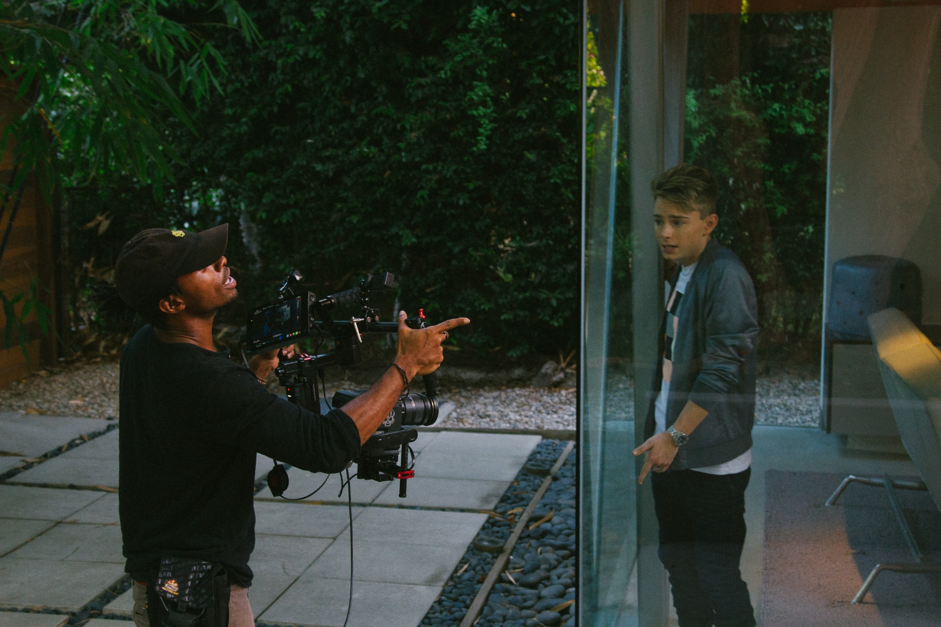 Q directing a young actor through a glass wall at dusk